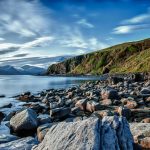 Serene coastal view with rocky shoreline and distant mountains under a vivid blue sky.